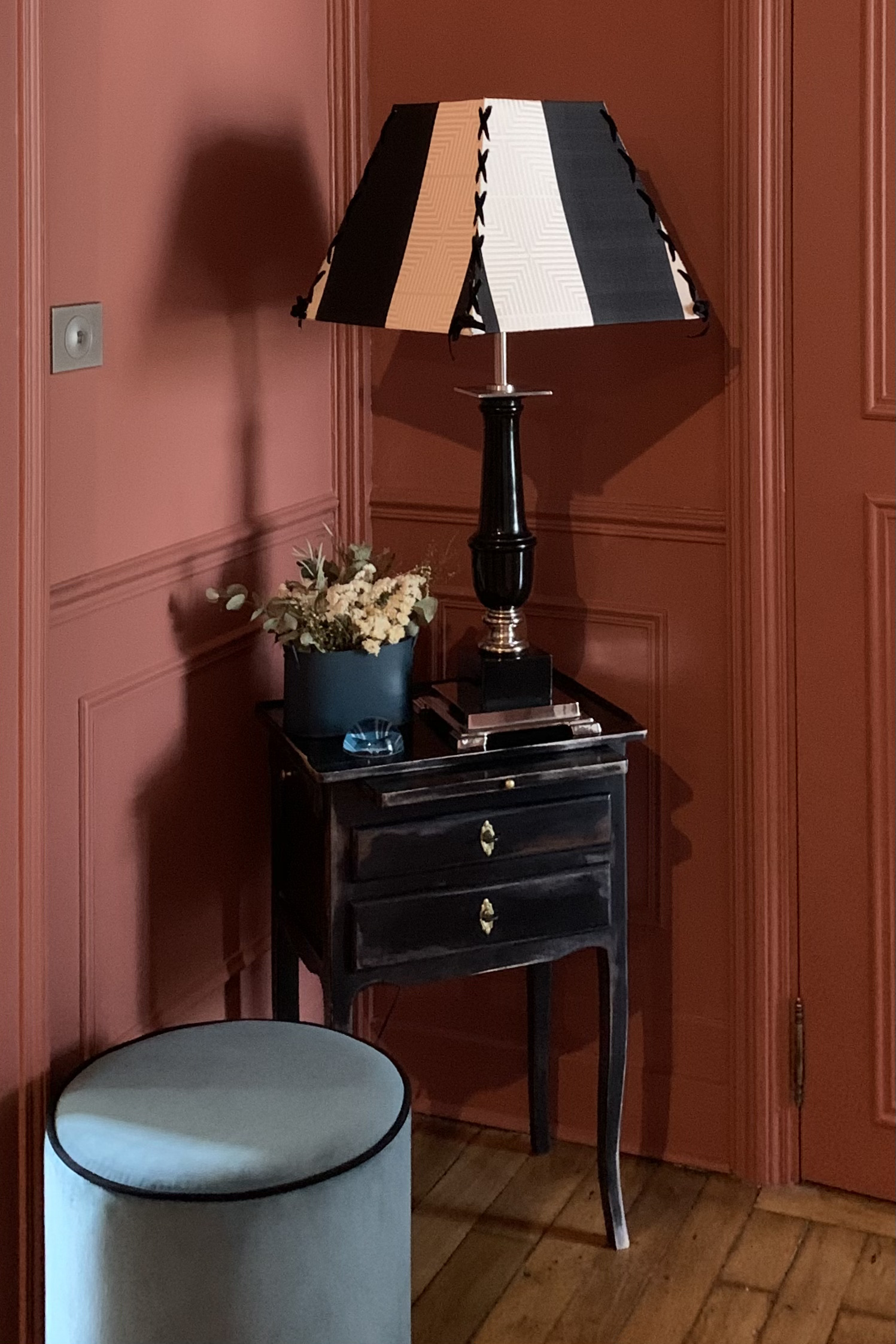 Projet Valérie - Entrée, mur rouge, meuble d'entrée noir avec une lampe et une fleur et un pouf bleu.