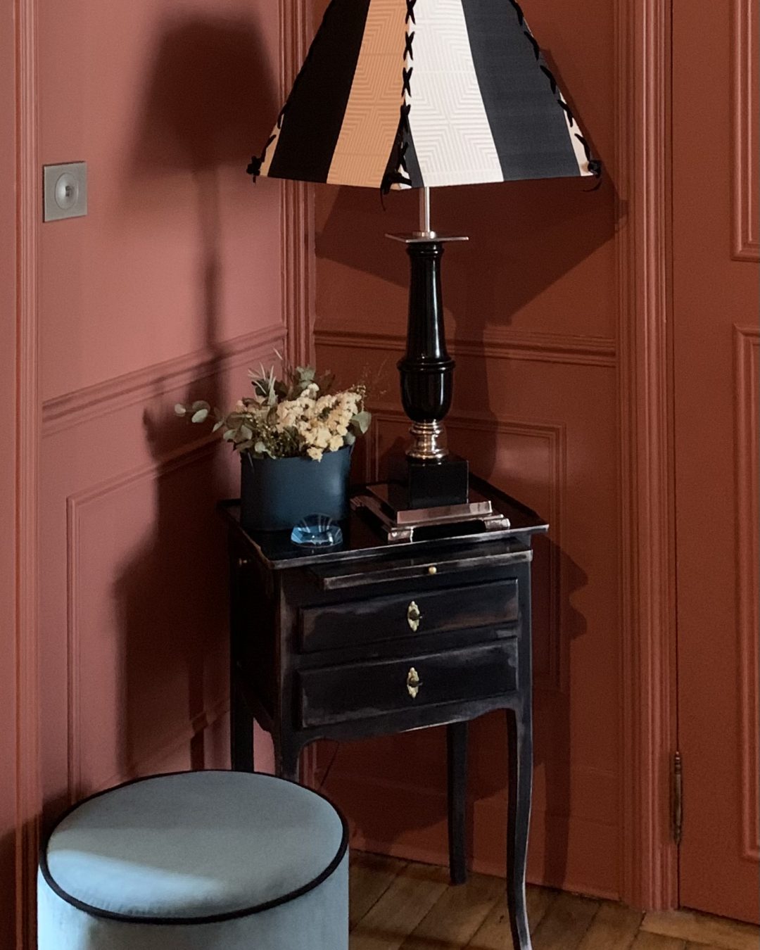 Projet Valérie - Entrée, mur rouge, meuble d'entrée noir avec une lampe et une fleur et un pouf bleu.